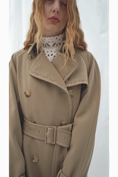 Stone trench coat with broad lapels traced in tiny eyelet cutwork. Self belt buckled wide at the waist. White high-neck underlayer punched with petal-shaped eyelets. First cold morning when the windows still sweat from summer.