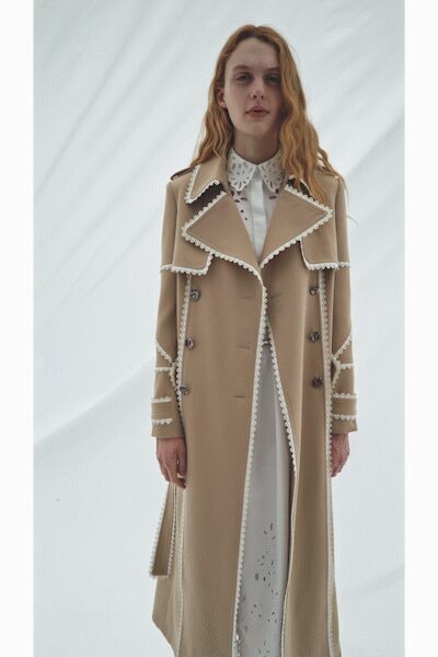 Long camel coat with oversized lapels edged in looped white trim. White eyelet dress falling straight beneath the open front. Pointed broderie anglaise collar laid over the coat neckline. Outside a winter chapel, just after the doors have opened.