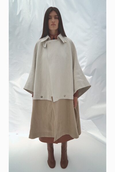 Ivory felted cape jacket with a skewed snap placket and wide bell sleeves. Oat linen shift dress falling in a blunt A-line below it. Brown leather boots, smooth and close through the ankle. Ferry terminal at first light, coffee cooling faster than expected.