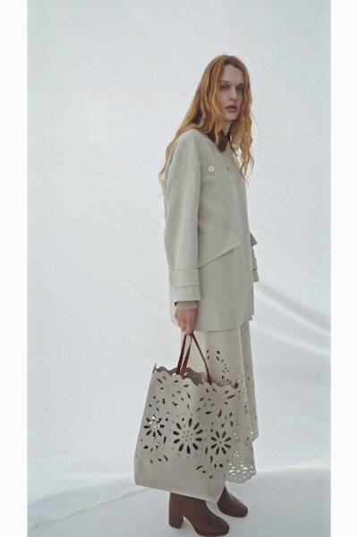 Pale collarless coat with broad cuffs and a slanted wrapped front. Matching midi dress, hem punched with petal cutwork over the calves. Oversized cream tote pierced with floral laser-cut holes. Chestnut leather ankle boots with blocky stacked heels. Crossing a museum atrium before the rain starts.
