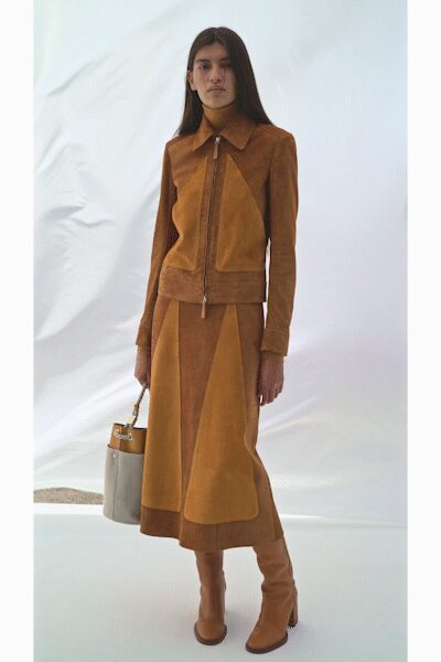 Pale beige knit turtleneck close to the throat. Caramel suede jacket pieced with ochre insets and a ring-pull zip. Midi suede skirt cut in long pointed panels to the hem. Cream bucket bag with a tan leather handle. Tan knee boots with a softly rounded toe. Dry grass underfoot, driving out to a low concrete house at dusk.