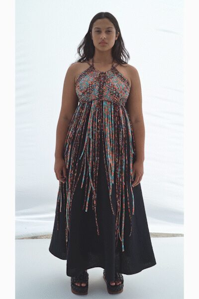 Halter-neck maxi with a densely ruched floral bodice and ribbon streamers falling from the waist. Black platform sandals with thick crisscross straps. Walking across a sun-bleached terrace after the wedding moved outdoors.
