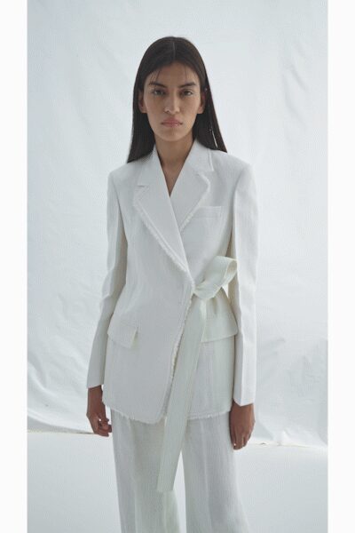 White wrap blazer with frayed edges and a long self-tie bow. Matching white plisse trousers, softly sheer below the blazer hem. Registry office at noon, flowers still in paper sleeves.