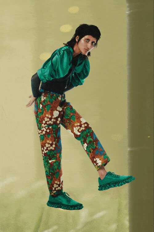 Jewel-green blouse with puffed sleeves gathered into a black corset waist. Brown jacquard trousers scattered with cream blossoms and curling green vines. Matching green slippers with a ruffled edge around the sole. Walking across a waxed hotel ballroom before the chairs are set.