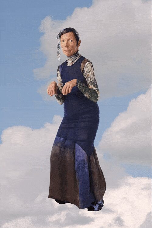 Pale papier-mache face mask tied with a dangling side ribbon. Second-skin rollneck in mottled grey-white marbled print. Ink knit column dress with dropped waist and sheer godet hem panels. Black shoes barely peeking under the pooling hem. Standing on a church roof while the weather turns strange.