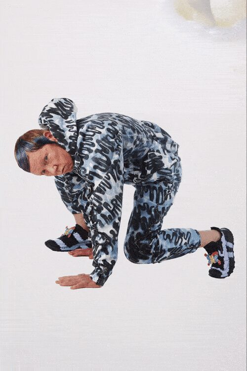 Grey velour hoodie covered in black graffiti loops, hood kicked upright. Matching velour joggers, dark spray-shadowing concentrated along the thighs. Black trainers with a spray of small bright charms at the laces. Crouching to fish dropped keys from under the laundromat dryer.