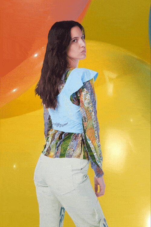 Pale blue knit shell with sharp ruffled cap sleeves and a scribbled hem. Printed long-sleeve blouse in watery vertical stripes, peplum gathered at the waist. Pale high-waisted trousers, smooth and slightly creased through the seat. Afternoon heat inside an inflatable maze at the fairgrounds.