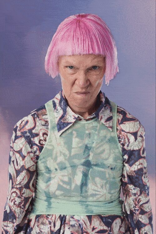 Bubblegum bob wig with blunt fringe cut straight across the brow. Swirled floral shirt in mauve and navy, pointed collar spread wide. Seafoam sheer apron top, bib front wrapped tight at the waist. At a craft fair table where the glaze on everything is still tacky.