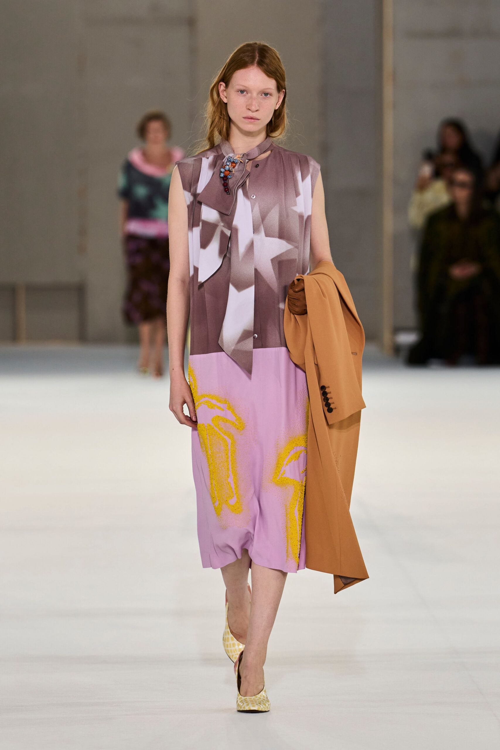 Sleeveless mauve dress with a scarf-tie neck and blurred white geometry. Clustered beads pinned at the throat, one drop hanging lower. Camel blazer carried folded over the arm, four dark cuff buttons. Butter yellow pumps with a crackled reptile-like finish. Walking back from a museum lunch with your jacket still over one arm.
