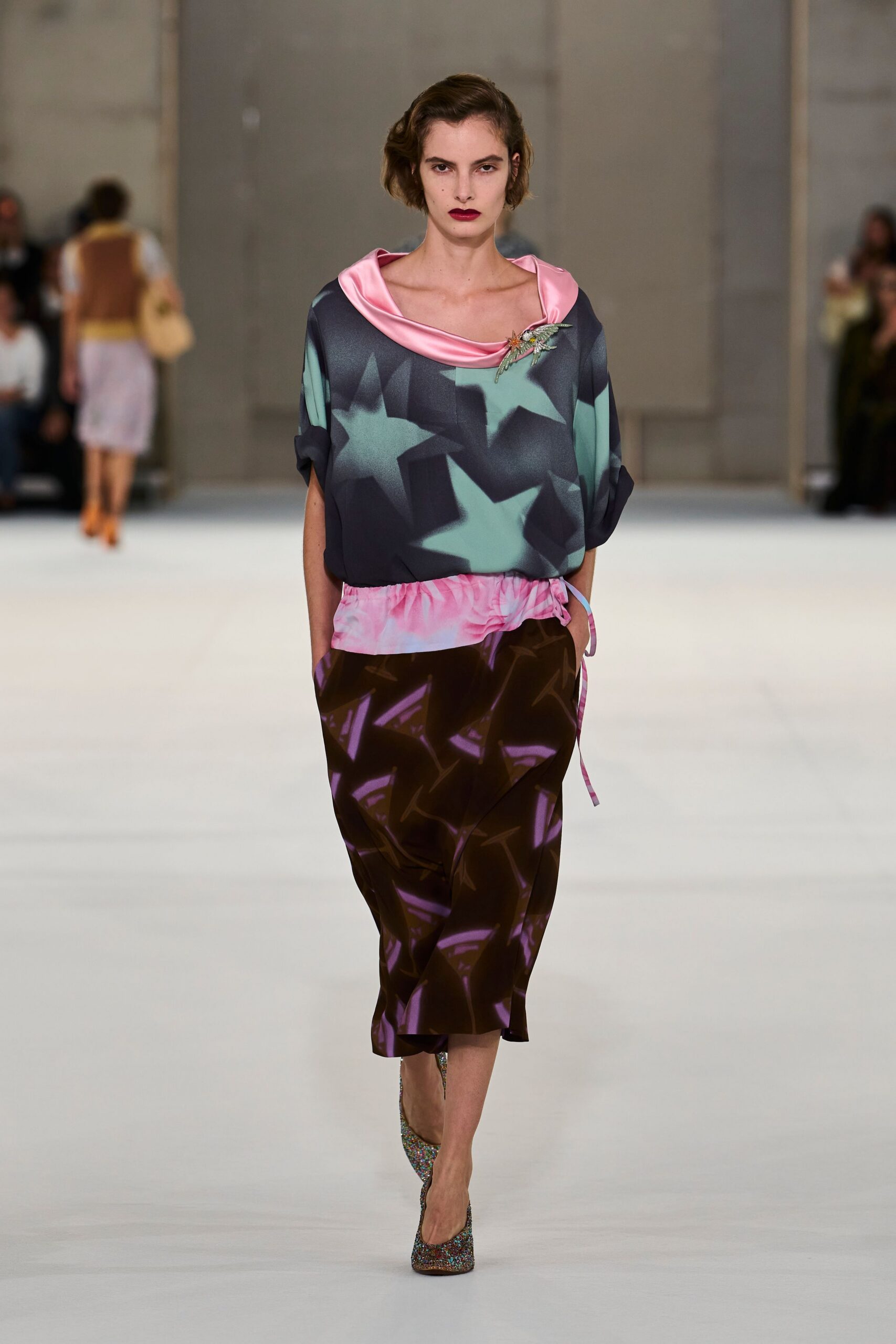 Charcoal satin top with mint star print and a pink rolled neckline. Beaded insect brooch pinned at the collarbone. Brown satin pencil skirt, pink drawstring waist hanging loose at one hip. Multicolored glitter pumps with a narrow pointed toe. Walking into a hotel bar before the wedding guests have come downstairs.