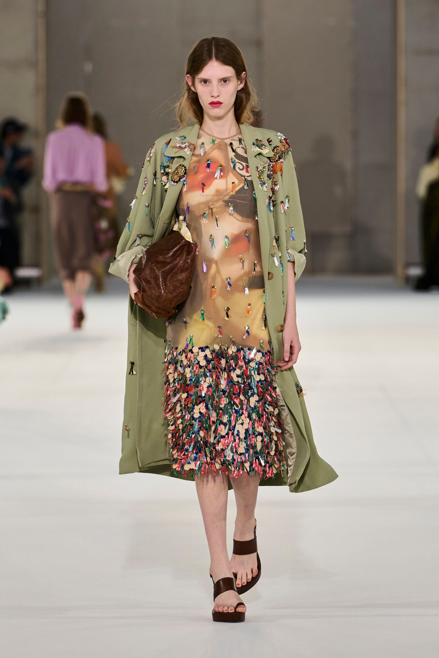 Sage trench strewn with tiny beaded doll charms and rolled cuffs. Sheer printed shift, hem drowned in dense paillettes and ribbon fringe. Crushed brown leather clutch tucked hard under one arm. Dark brown platform sandals with a wide ankle strap. Walking into a hotel atrium after buying trinkets from every museum shop.