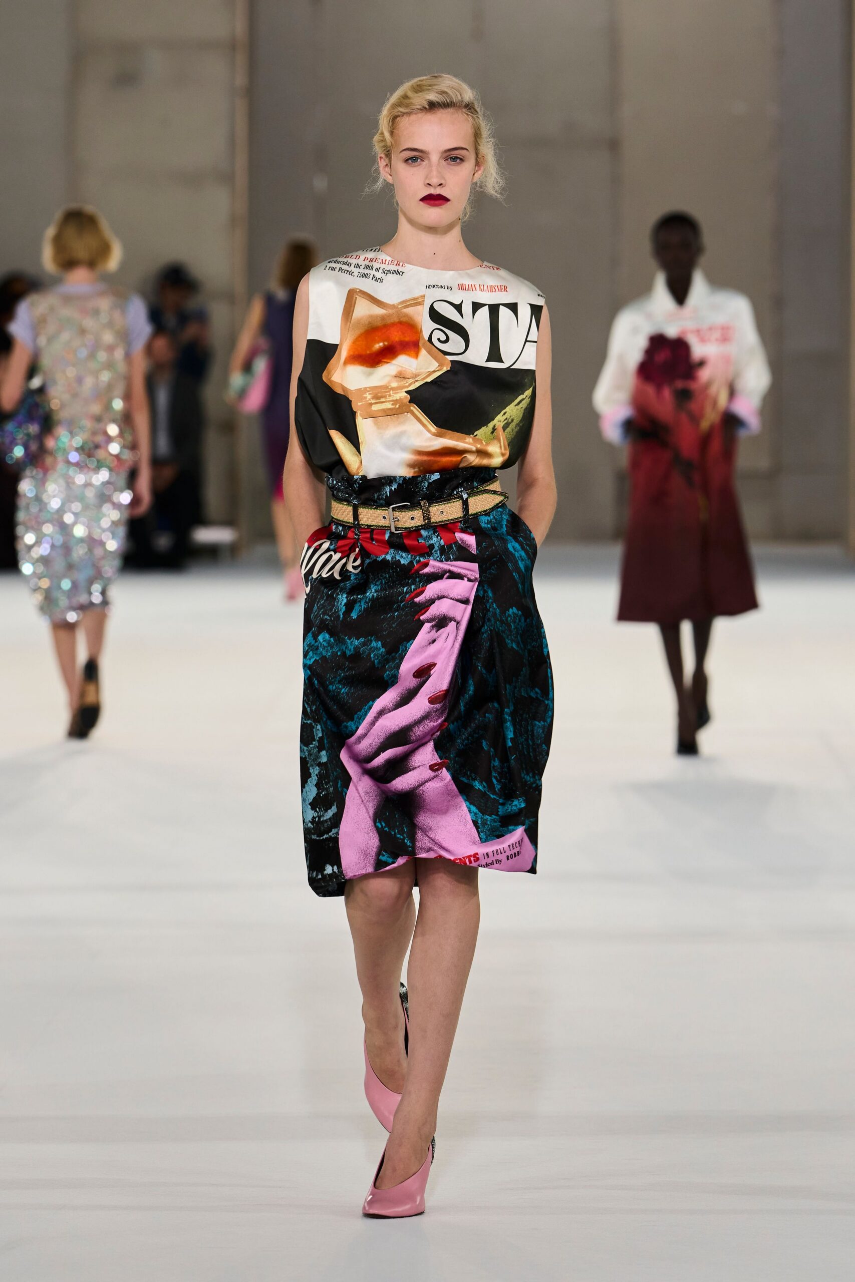 Sleeveless satin shell printed like a torn magazine cover. Wide beige snakeskin belt with a square silver buckle. Knee-length satin skirt, pink hand graphic wrapped across the front. Bubblegum patent pumps. Leaving a print studio with ink still under one thumbnail.