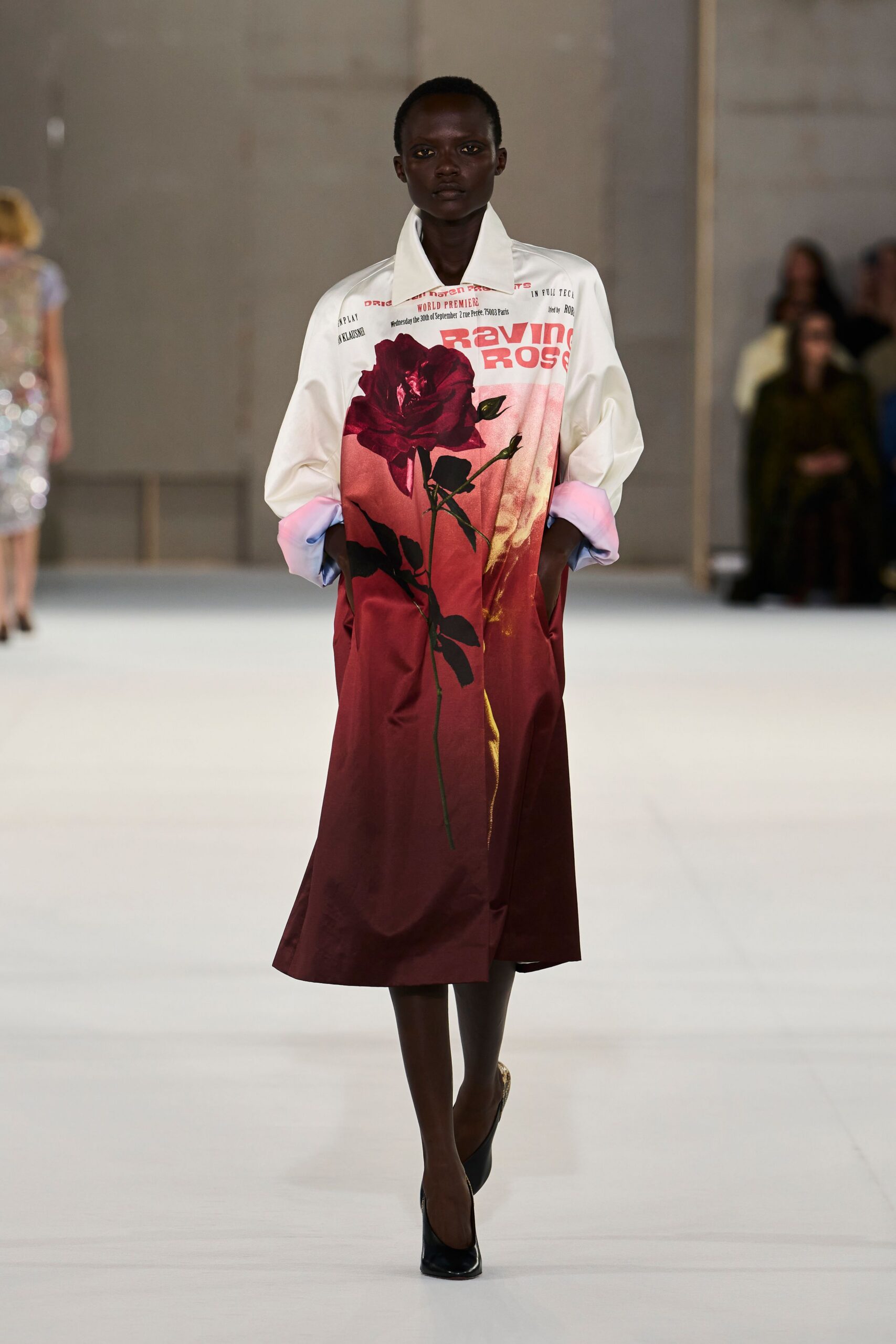 Glossy white shirting printed with tabloid text and a single oversized crimson rose. Turned-back cuffs washed from pink into pale blue. Black pointed pumps, cut clean and bare at the instep. Crossing a museum atrium with a bouquet wrapped in yesterday's paper.