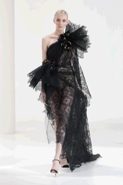 One-shoulder black lace gown with a sheer train and scalloped hem. Exploded tulle ruffle climbing off the bare shoulder. Dark floral clusters pinned at the neckline and wrist. Black ankle-strap sandals with a pale platform peeking forward. Whatever arrives overdressed to a winter gala and stays till chairs go up.