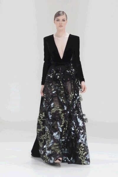 Black velvet bodice with a plunge almost to the waist. Floor-length black skirt crusted in oversized paillettes and sheer tulle panels. Black open toe sandals peeking beneath the hem. Crossing a hotel ballroom after midnight, sequins catching the spill from the bar.