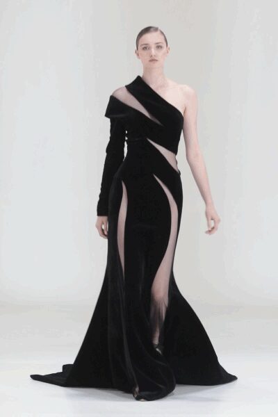 Black velvet one-sleeve gown slashed into curved nude cutouts. Floor-pooling back panels spreading behind the hem. Black pointed toes just breaking the front slit. Museum atrium after hours, every footstep carrying farther than expected.