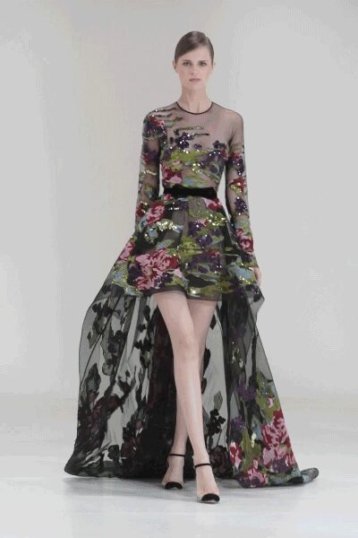 Sheer black tulle gown densely embroidered with sequined painterly florals. Black satin band cinching the dropped waist seam. Black ankle-strap heels with a peep toe. Crossing a marble foyer after midnight with petals stuck to the hem.
