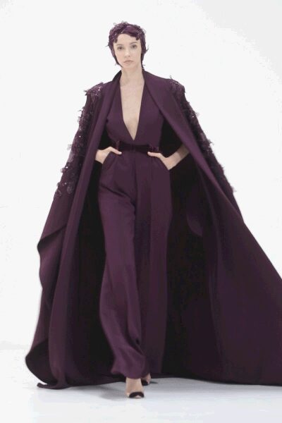 Plum sequined skullcap fitted close over the cropped hair. Floor-sweeping aubergine cape, shoulders crusted with tonal paillettes. Deep-plunge aubergine crepe jumpsuit, waist cinched and legs falling loose. Black pointed heels. Crossing a museum atrium after hours, echo following a hem this long.