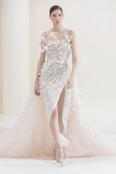 Strapless blush tulle gown densely beaded in radiating lines, thigh slit opening through the drape. Sheer overskirt strewn with petal appliques, one shoulder drifting into a floral spray. Satin platforms under a gauzy hem. For crossing a hotel ballroom after the vows, champagne already warm.
