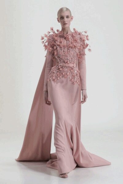 Blush column gown with beadwork and clustered petal appliqué erupting at bodice and shoulders. Long blush cape falling clean from the shoulders to a pooled train. Peep-toe heels barely visible under the hem. For the first slow turn in a ballroom gone quiet after midnight.