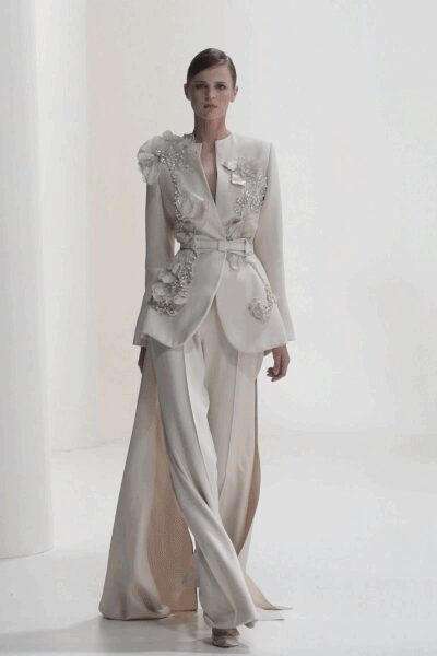 Ivory satin blazer with crystal-beaded blossoms climbing the lapel and hip. Self-tie sash cinching the waist in a soft knot. Fluid ivory trousers split open at the outer leg into long draped panels. Pale pointed heels barely visible under the hem. For signing papers in a room with too much echo.