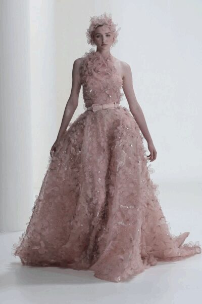 Blush one-shoulder ball gown buried in petal appliqué and scattered sequins. Wide self belt, tied flat at the waist. Matching pink floral tuft pinned into clouded hair. At the first fitting after the invitation says black tie only.
