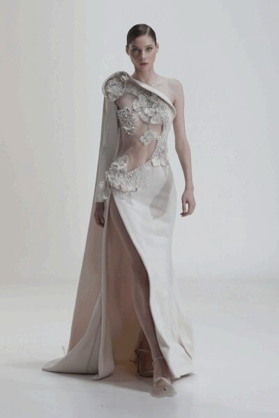 One-sleeved ivory satin gown with a molded shoulder and thigh-high split. Sheer nude tulle cutout strewn with pearly floral embroidery and crystals. Nude mesh pumps with pointed toes. Registry-office stairs, bouquet paper still crackling in one hand.