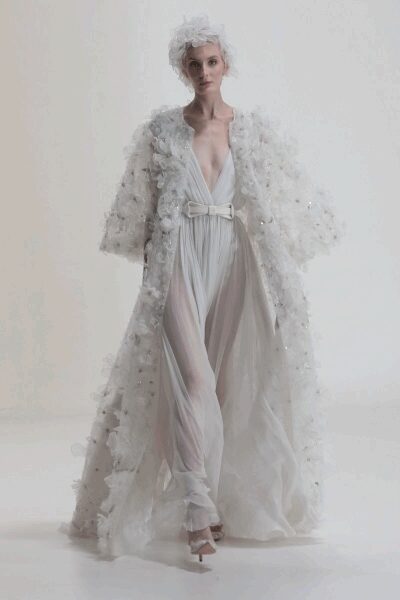 Wispy white tulle headpiece, feathered into an uneven cloud. Floor-length ivory coat crusted with organza florets and scattered bead shine. Plunging white chiffon gown, softly pleated from a wide buckled waist. Pointed ivory heels peeking beneath the froth. Crossing a hotel ballroom after the wedding cake has already been cut.