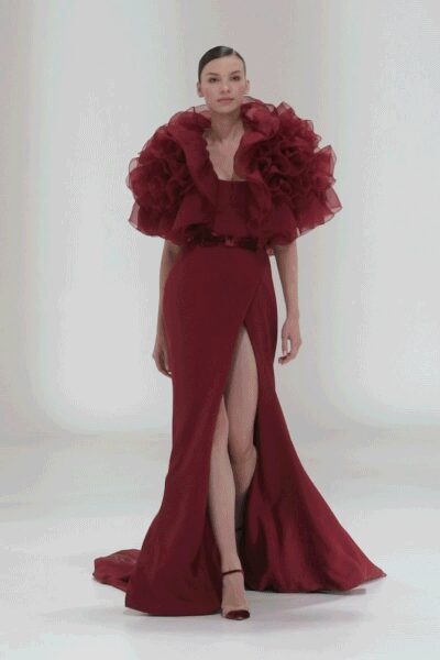 Burgundy column gown with a thigh-high split and pooled satin train. Cloud of oxblood organza ruffles swallowing the shoulders and upper arms. Narrow dark belt pinching the waist. Barely-there ankle-strap heels. For entering a ballroom after everyone has already taken their seats.
