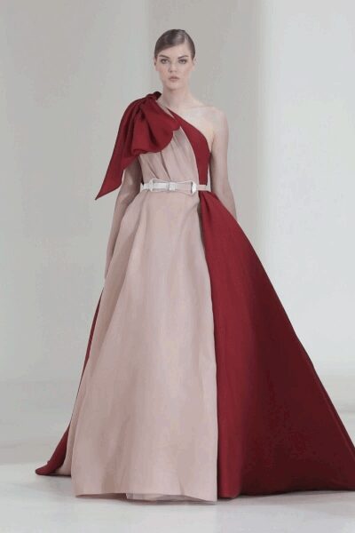 One-shouldered blush column gown split by a oxblood panel and train. Oversized oxblood bow collapsing over one shoulder in crisp folds. Slim white belt, squared buckle catching the waist high. For arriving first at a winter wedding in an echoing civic hall.