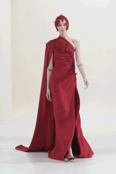 Red floral headpiece cupping the head close like clustered petals. One-shoulder crimson column gown with beaded appliqué climbing the bodice. Floor-pooling cape falling from the left shoulder in heavy satin folds. At the charity gala after the speeches, crossing the room too late to hide.