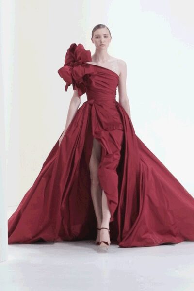 One-shoulder crimson taffeta gown, bodice ruched tight with a giant bow at the shoulder. Voluminous train opening from a wrapped front panel to show one leg. Barely there ankle-strap heels. Crossing an echoing hotel corridor just before the ballroom doors open.
