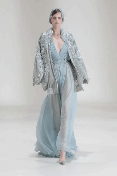 Silver sequin hood cupping the head close. Oversized silver paillette coat, sleeves ballooned and hanging open. Ice-blue chiffon gown with a plunging twist-front bodice and trailing panels. Clear pointed heels. Crossing a hotel ballroom after the chairs have been stacked.