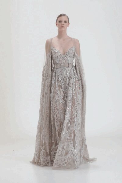 Blush tulle gown densely embroidered in silver scrolls and scattered crystals. Floor-length sheer cape sleeves falling from the shoulders. Narrow metallic belt fastening the high waist. For the staircase descent before the room goes quiet.