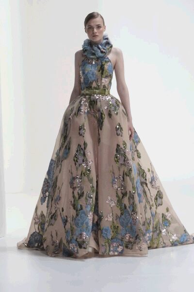 Blush ball gown scattered with blue floral sequins and a moss-green waist bow. Cloudy blue ruffle wrapped thick at the throat. Museum fundraiser, standing under the last quiet chandelier.