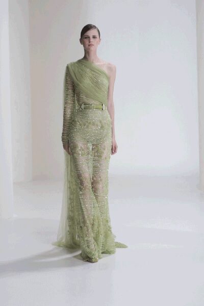 Celadon one-sleeve net gown drenched in sequins, with a sheer train. Narrow matching belt, small rectangular buckle catching at the waist. Pointed shoes veiled under the same glittering mesh. Crossing a hotel atrium before sunrise, everything still smelling faintly of lilies.