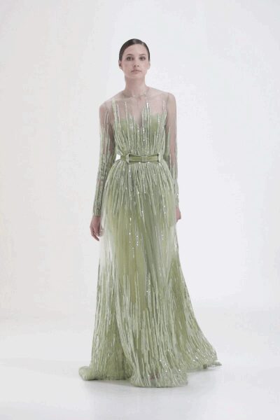 Pale pistachio column gown under clear sequined fringe, plunging V barely held. Matching satin belt knotted at the waist with short trailing ends. Crossing a hotel atrium at dawn after the flowers have been cleared.