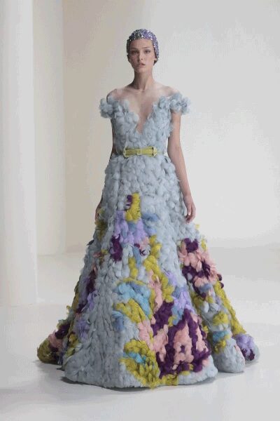 Silver beaded cap fitted close over the head. Ice-blue gown built from tufted cloudlike clusters, plunging at the front. Pale chartreuse belt cinching the waist. Skirt erupts into lilac, butter and violet floral mounds at the hem. Crossing a hotel ballroom carpet after the florists have already left.