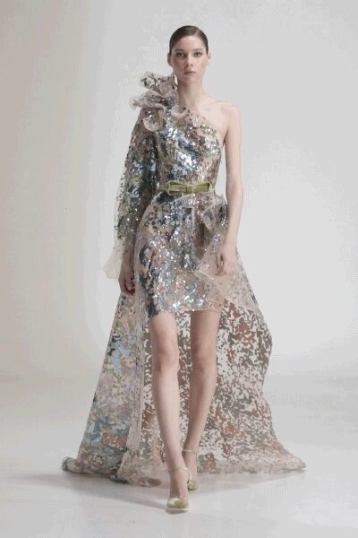 One-sleeve sequin mini in watery silver, shoulder exploding into organza ruffles. Slim olive belt cinching the waist. Sheer floral train spilling from the back in mottled lace. Pale ankle-strap heels. For the first dance after the garden lights finally come on.