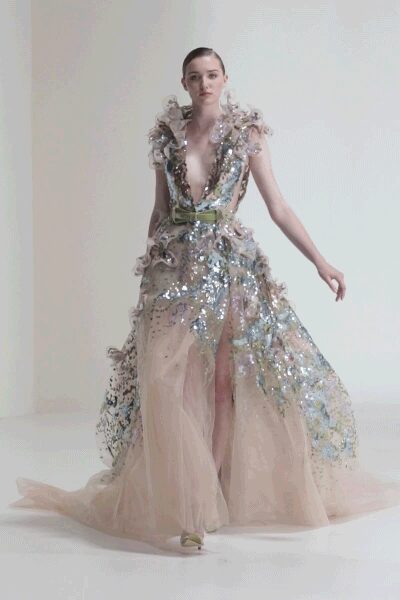 Nude tulle gown crusted with iridescent paillettes, plunging to the waist. Organza frills rising at the shoulders like crushed petals. Pale green satin belt breaking the shimmer at the middle. Pale peep-toe heels. Crossing a hotel ballroom after the orchestra has packed away.
