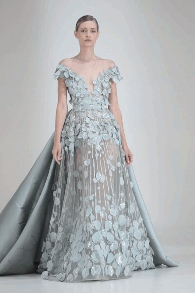 Ice-blue tulle gown with off-shoulder petal straps and plunging illusion V. Layered leaf paillettes scattered dense at the hem, trailing up in strands. Sculpted satin train flaring from the back like folded wings. For standing at the top of a staircase after everyone else has gone down.