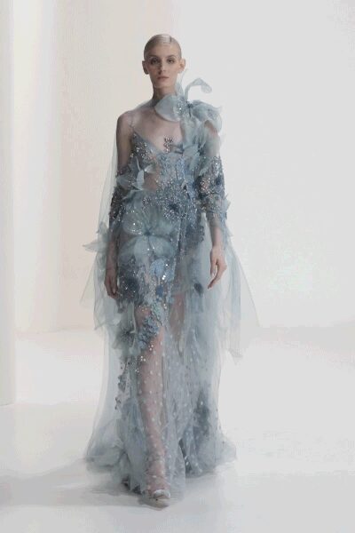 Ice-blue tulle gown crusted with sequins and appliqued blossoms. Sheer draped cape sleeves falling from the shoulders in long panels. High organza ruffle curling up one side of the neck. Silver barely-there sandals. Crossing a hotel lobby at dawn with petals caught in the train.