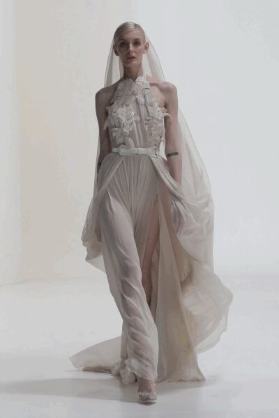 Floor-length tulle veil falling from a close skullcap. Blush halter gown with ruched chiffon trousers and appliqued flowers at the bib. Narrow self belt cinching the dropped waist. Pale peep-toe heels under the pooling hem. City hall atrium, after the ceremony but before anyone found the flowers.