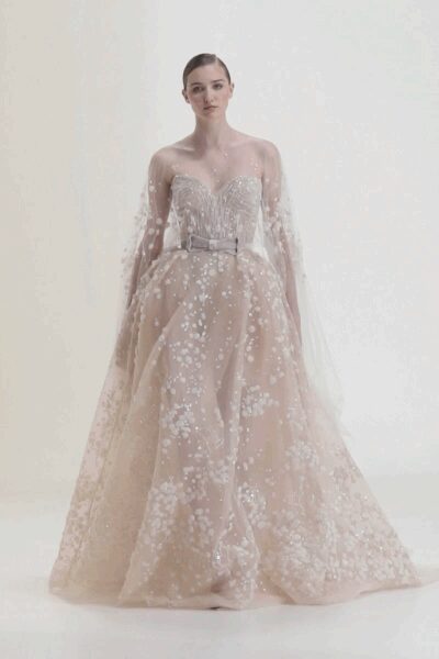Sheer tulle cape scattered with white floral applique, falling from bare shoulders. Blush strapless ballgown with a sweetheart bodice and soft glittered underlayer. Narrow silver belt, centered with a small rectangular buckle. For the first slow turn in a fitting room full of pins.