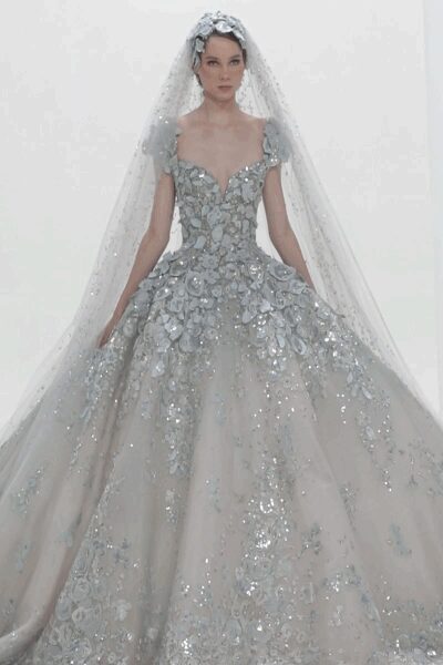 Cathedral veil dusted with sequins, topped by clustered fabric blossoms. Ice-blue ball gown crusted in appliqued flowers and silver beading, skirt billowing from a dropped waist. At the chapel door just before the organ starts.