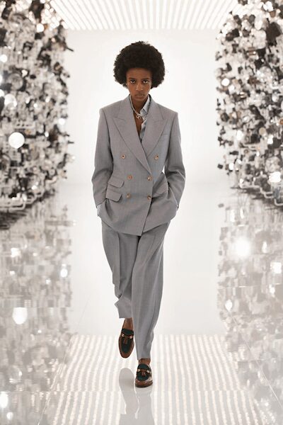 Pale grey double-breasted blazer with peaked lapels and low flap pockets. Chunky silver chain tucked into the open throat. Loose straight trousers in matching suiting cloth, lightly breaking at the loafer. Dark tassel loafers with a green apron seam. Walking a bright hotel corridor before anyone else is up.