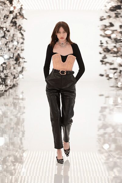 Black long-sleeve bodysuit with a deep scooped neckline. Blush corset bustier, topstitched panels curving over the bodysuit. Black leather trousers slouched at the hips with pleats under a logo buckle. Leopard pointed heels with slim ankle straps. Whatever you wear home when the penthouse party spills into daylight.