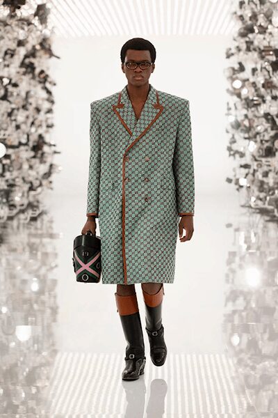 Square black acetate frames. Mint double-breasted coat in tiny geometric jacquard, tan piping tracing the lapels. Cylindrical black holdall with pink and mint chevrons. Black leather knee boots with tan cuffed shafts and silver buckles. Walking into a grand hotel after the porter has already taken the trunk.