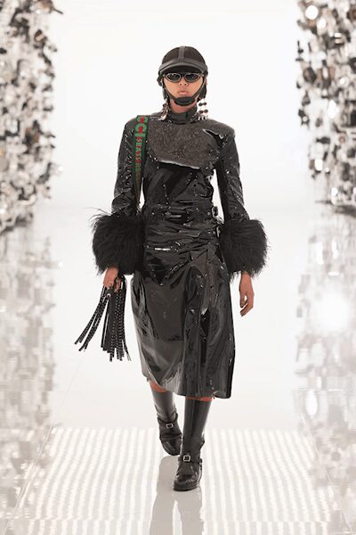 Black leather cap with ear-covering straps fastened under the chin. Dark oval lenses in a pale frame. Black patent dress with a high neck and shaggy fur cuffs. Fringed black handbag swinging from a short handle. Knee-high black boots with a strap across the instep. Crossing a frozen plaza with shopping still to finish.