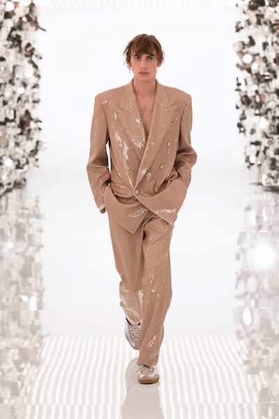 Blush double-breasted blazer washed in scattered sequins, cut with broad shoulders. Matching straight trousers, sequins thinning toward the cuff break. Pale low-top sneakers with thick laces and rubbery soles. Crossing a hotel atrium at noon after sleeping in yesterday's makeup.